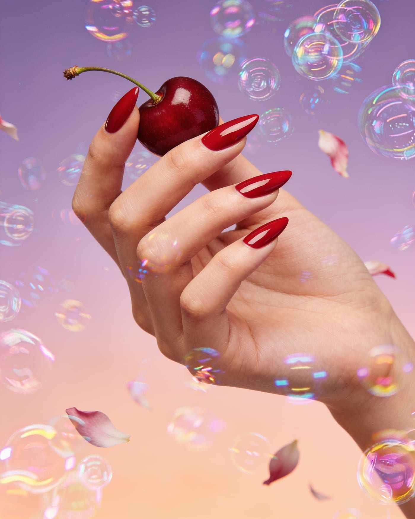 Long almond nails in glossy cherry red, holding a cherry by its stem among floating bubbles.