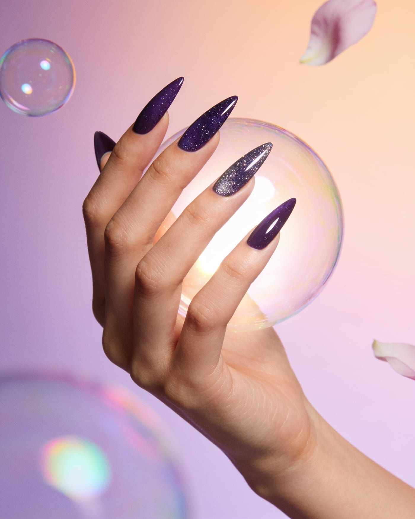 Long stiletto nails in deep plum, two of them sparkling with glitter, a hand cradling a soap bubble.